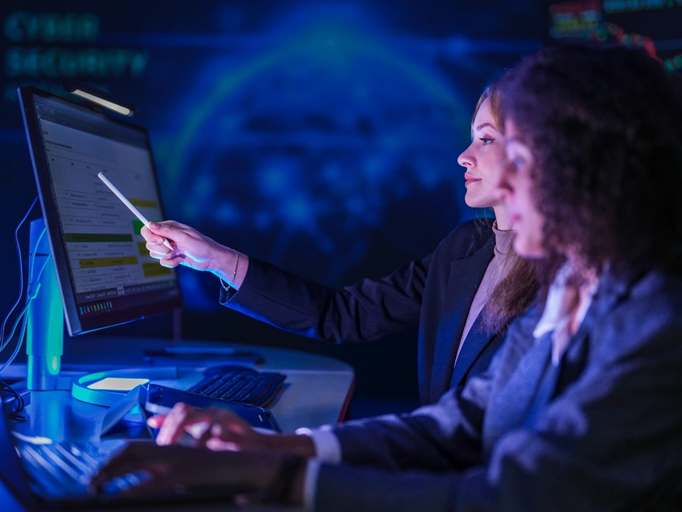 Cyber security specialists monitoring global network threats. Female tech expert pointing at digital system data on screen while partner analyzing code at night.