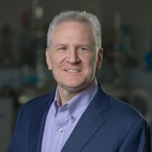 A man wearing a navy suit jacket and light purple shirt is photographed in an industrial environment.