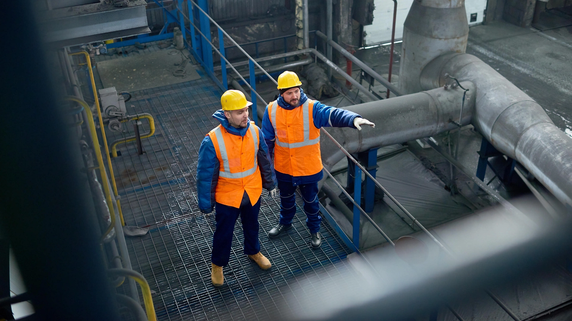 High angle view of bearded engineers wearing uniform and protective helmets standing at spacious production department of modern plant and brainstorming on joint work, High angle view of bearded engineers wearing uniform and protective helmets standing at spacious production department of modern plant and brainstorming on joint work