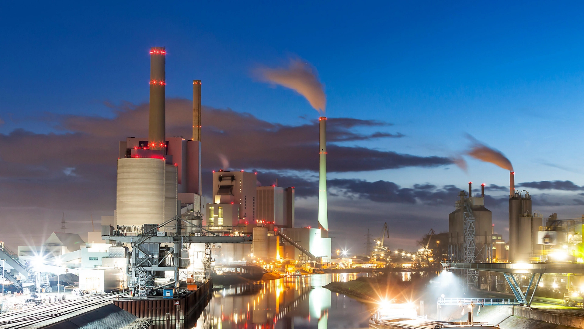 A coal power plant sits at the river's edge at dusk.  Lights from the buildings and streets glow, and a reflection of the whole scene can be seen on the water of the calm river below.