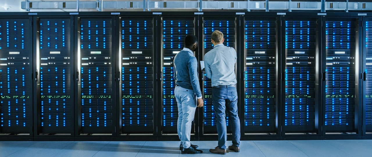 Bearded IT Technician in Glasses with a Laptop Computer and Black Male Engineer Colleague are Talking in Data Center while Working Next to Server Racks. Running Diagnostics or Doing Maintenance Work.