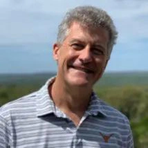 A man is seen standing outdoors against a backdrop of greenery and a blue sky.