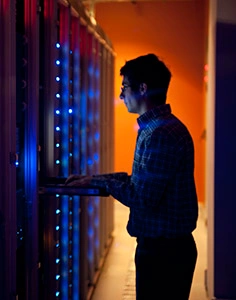 The interior of a modern server room in a data center.  An IT engineer is busy configuring the servers. The room is dark, but the servers themselves are lit.  The servers at the left side are lit in blue, while the ones at the other end are lit by an orange light source coming from the right side.